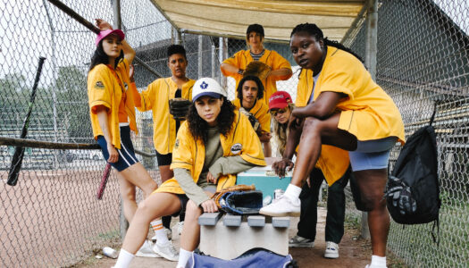 Group of diverse softball players in yellow jerseys posing in a dugout behind a chain-link fence, with gloves and gear nearby.
