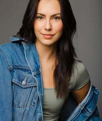 alex-hook – LezWatch.TV Portrait of a woman with long dark hair, wearing a denim jacket over a green top, smiling softly at the camera against a neutral gray backdrop
