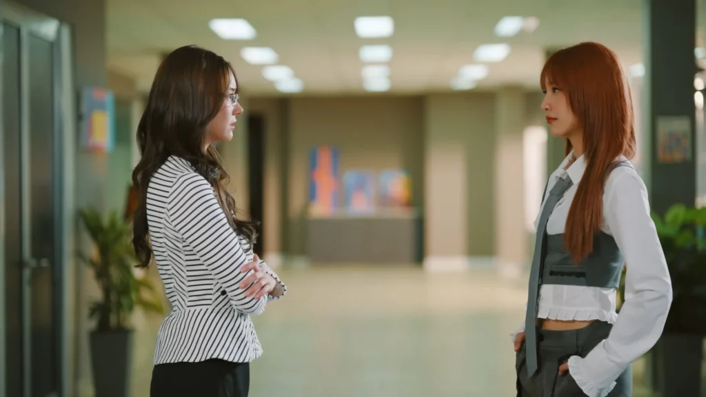 Two women face each other in a hallway, one wearing a striped top with crossed arms and glasses, the other in a white shirt with a gray vest.
