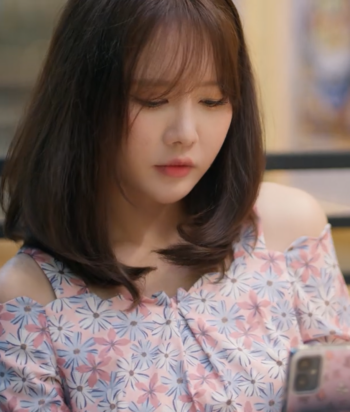 Woman with shoulder-length brown hair looking at her smartphone, pastel pink floral off-shoulder top worn outdoors/indoors.