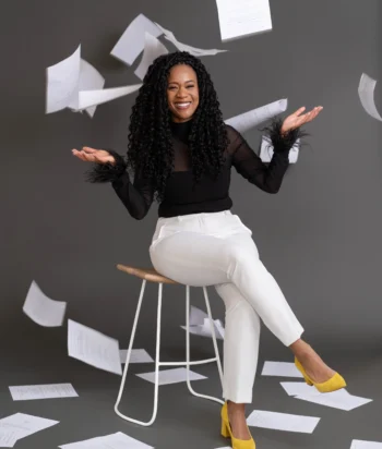 129_Waldenjpg – LezWatch.TV Smiling woman with curly hair sits on a stool as papers fly around her in the air against a gray background.