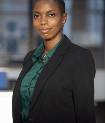 Professional woman in a green blouse and black blazer posing for a headshot in an office setting.