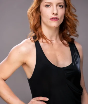 Afton_Rentz – LezWatch.TV Portrait of a woman with red hair wearing a black sleeveless top, standing with one hand on her hip against a gray background, looking at the camera.