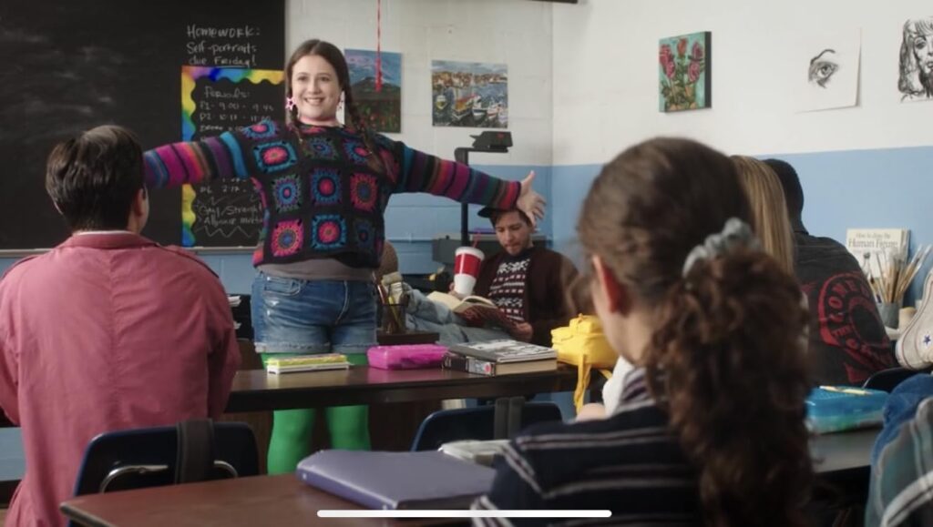 Teacher with arms outstretched stands at the front of a colorful classroom as students sit at tables and listen.