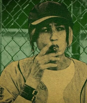 Person wearing a cap making a shushing gesture with a finger to their lips, in front of a chain-link fence.