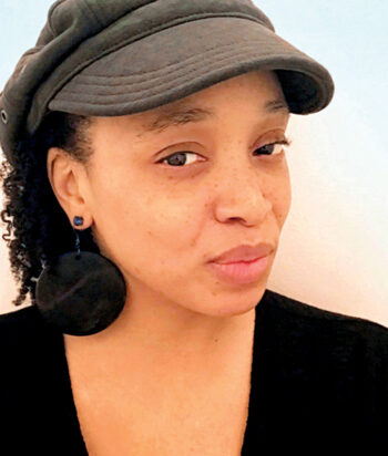 Cenophia – LezWatch.TV Portrait of a woman wearing a brown cap, black top, and large black circular earrings against a light background.