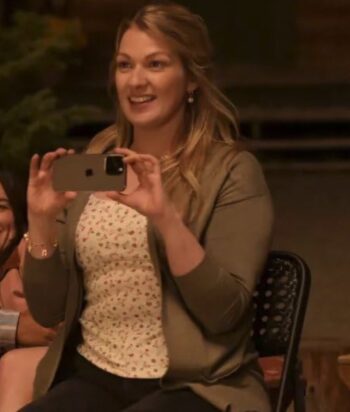 Woman seated, smiling, holds a smartphone to take a photo or video at an event.