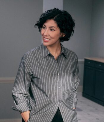 Didi – LezWatch.TV Woman with short curly black hair in a gray striped button-down shirt, smiling and looking to the left in an office setting.
