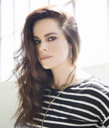 Emily-Hampshire – LezWatch.TV Portrait of a woman with long wavy brown hair, wearing a black-and-white striped top and layered gold necklaces, looking at the camera.