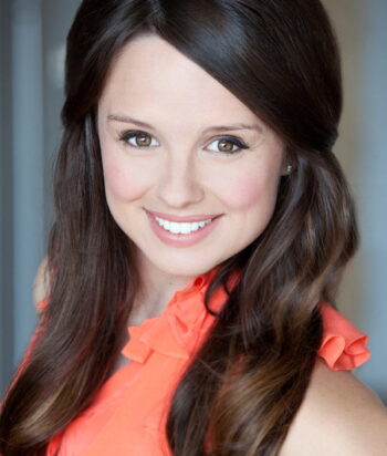 Emma-Hunter – LezWatch.TV Smiling woman with long dark hair wearing an orange blouse or top, facing the camera in a close portrait liberty style.