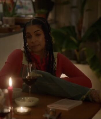 Woman with braided hair in a red sweater sits at a dining table, a glass of red wine and a candle nearby, with plants in the background.
