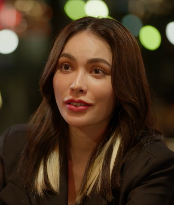Irene – LezWatch.TV Woman with dark hair and blonde highlights speaks at a dimly lit venue, wearing a black blazer.