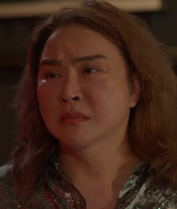 Close-up of a distressed woman with shoulder-length red hair wearing a glittering blouse in a dim setting.