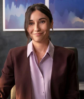 Smiling woman in a burgundy blazer and lavender blouse, seated at a desk in an office with blue abstract art behind her.