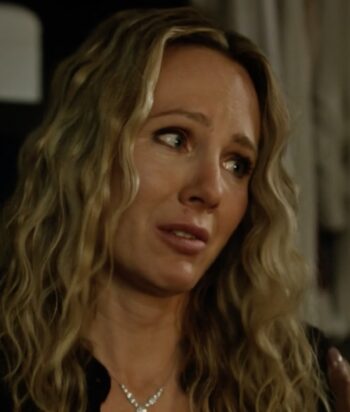 Close-up of a woman with long wavy blonde hair looking to the right with a worried expression in dim lighting.