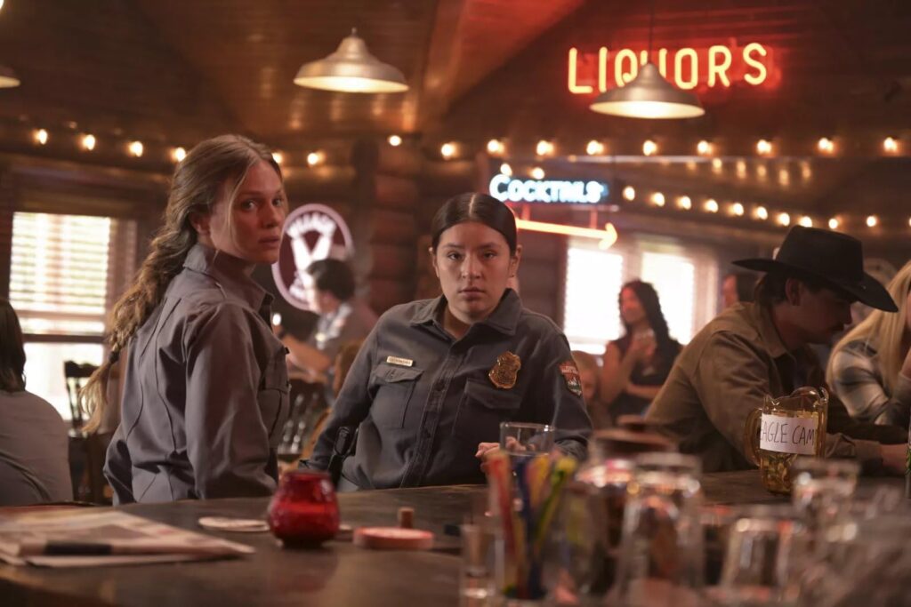 Two women in gray uniforms stand at a dimly lit bar, looking toward the camera, with a neon LIQUORS sign in the background.