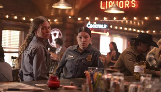 Two women in gray uniforms stand at a dimly lit bar, looking toward the camera, with a neon LIQUORS sign in the background.