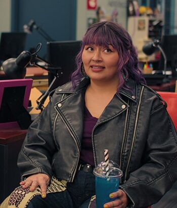 Millie-Maktana – LezWatch.TV Woman with purple hair wearing a black leather jacket sits in a coworking space, holding a blue beverage with a striped straw.