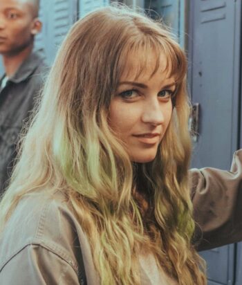 Screenshot – LezWatch.TV Young woman with long wavy blonde hair posing near blue lockers, looking at the camera.