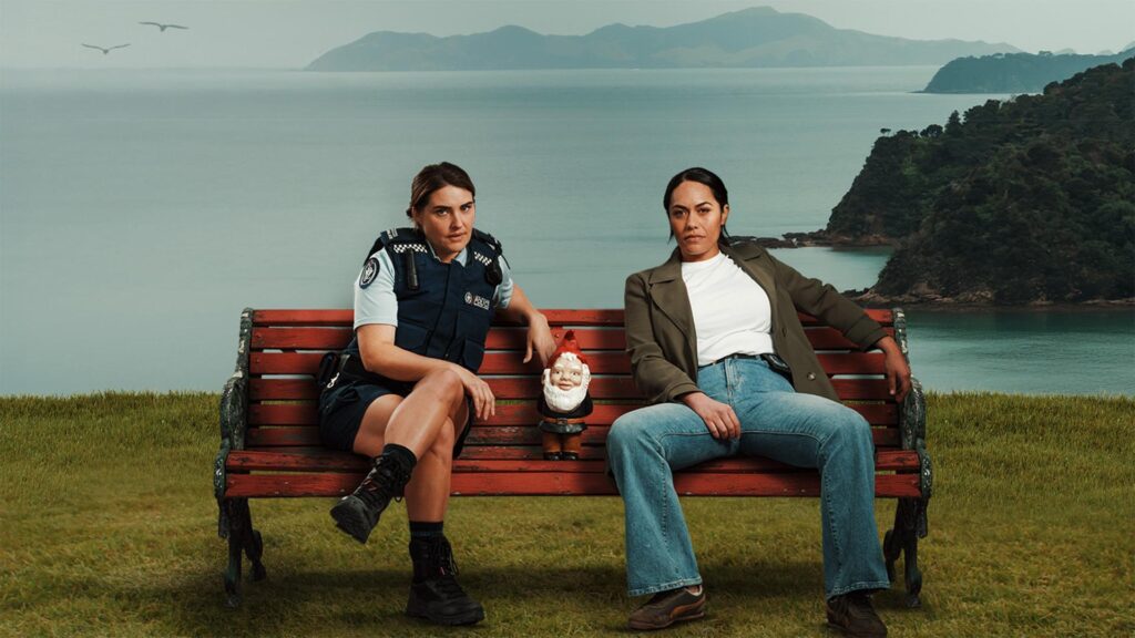 Two women sitting on a red bench by the water; one in police uniform, the other in a jacket, with a garden gnome between them.