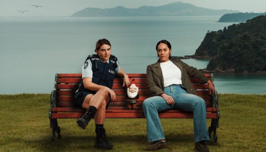 Two women sitting on a red bench by the water; one in police uniform, the other in a jacket, with a garden gnome between them.