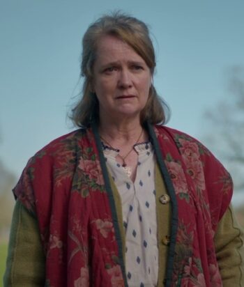 Older woman standing outdoors in a red floral shawl over a light blouse, looking thoughtful.