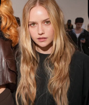 Amelie-Hoeferle – LezWatch.TV Young woman with long wavy blonde hair wearing a black top, looking at the camera in a backstage fashion show setting.
