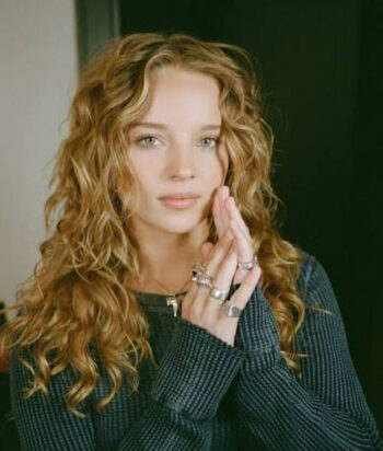 Bo-Bragason – LezWatch.TV Woman with curly blonde hair and multiple rings, wearing a dark sweater, hands pressed together near her face.