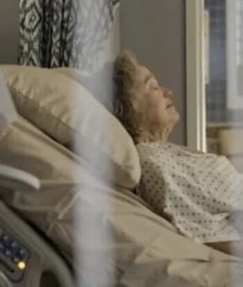 Elderly woman resting in a hospital bed, head propped on pillows, wearing a patient gown near a window with curtains in the background.