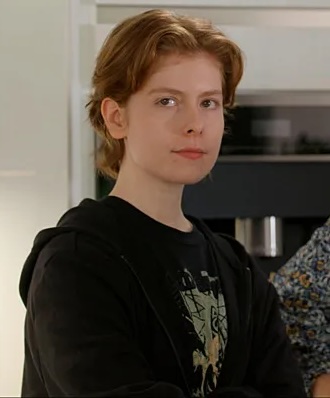 Young woman with short blond hair wearing a black hoodie, standing in a modern kitchen area.