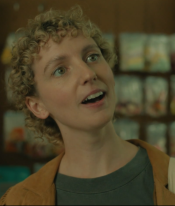 Woman with short curly blonde hair looking up with a surprised expression in a shop, wearing a green shirt and tan jacket with a tote bag over her shoulder.