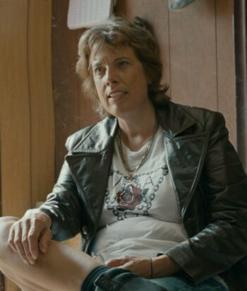 Person with short light brown hair in a black leather jacket and graphic tee, seated indoors near a wooden wall.
