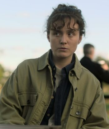 Young woman wearing a khaki jacket, looking to the left against a blurred park background.