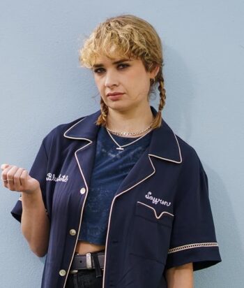 Young woman with blonde braided hair wearing a navy short-sleeve jacket over a blue top, standing against a pale blue wall.