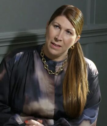 Woman with long brown hair in a ponytail, wearing a dark tie-dye blouse and chunky gold necklace, speaking in a studio setting.