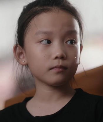 Young girl with dark hair in a black shirt sits, looking to her left with a thoughtful expression.