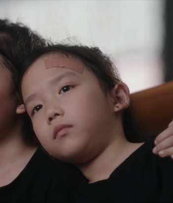 Young child resting against an adult with a bandage on the forehead, looking somber.